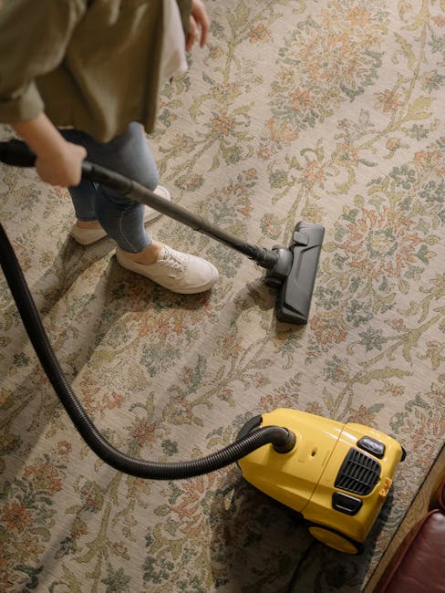Close-up of a home carpet cleaning vacuum cleaner with a transparent water tank filled with water and bubbles, connected to a flexible hose leading to a black rectangular floor attachment on a pinkish