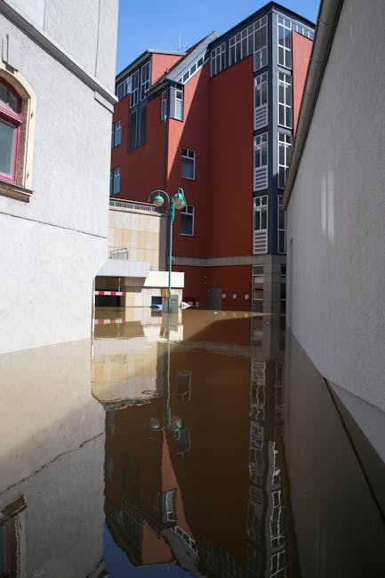 A street scene in Cambridge Heath shows a flood-affected area with muddy water covering the ground and partially submerging the lower parts of surrounding buildings. The image features a modern, multi