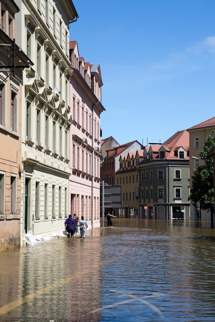 A street scene in Cambridge Heath shows a flood-affected area with muddy water covering the ground and partially submerging the lower parts of surrounding buildings. The image features a modern, multi
